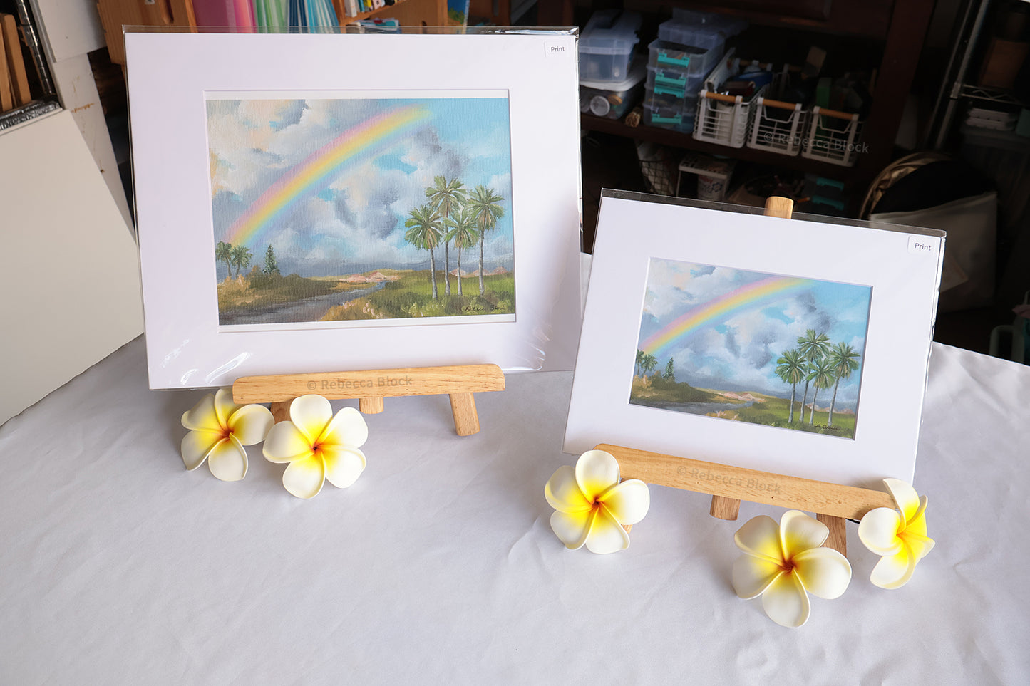 Two matted landscape art prints showing a rainbow over palm trees, displayed on wooden easels on a white tablecloth with plumeria flowers in front.