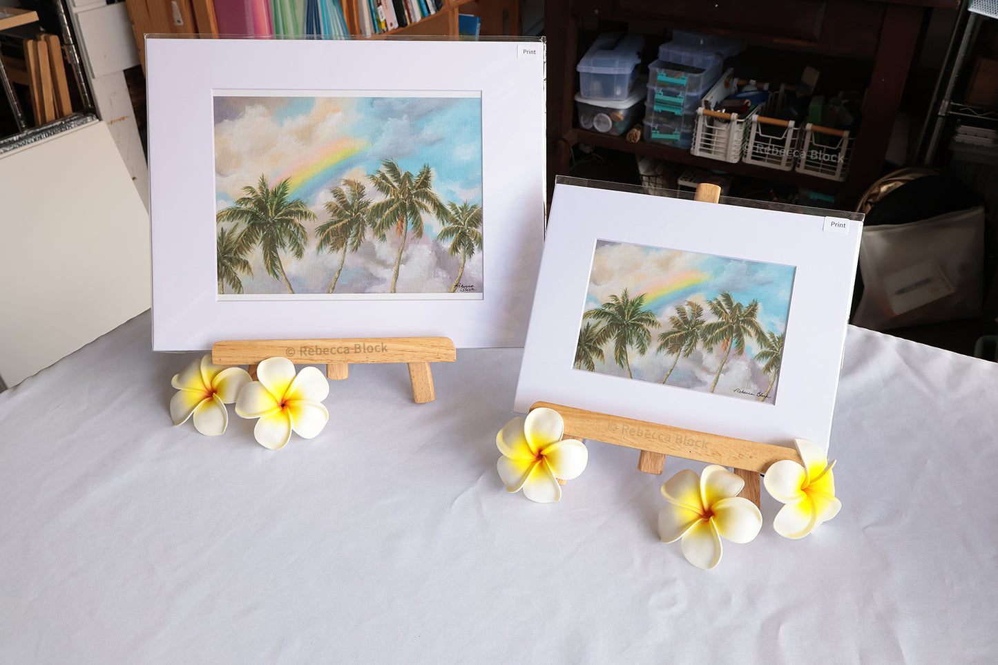 Two matted art prints showing tropical palm trees with a rainbow, displayed on small wooden easels on a white tablecloth, with plumeria flowers placed in front.