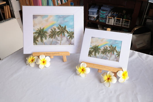 Two matted art prints showing tropical palm trees with a rainbow, displayed on small wooden easels on a white tablecloth, with plumeria flowers placed in front.