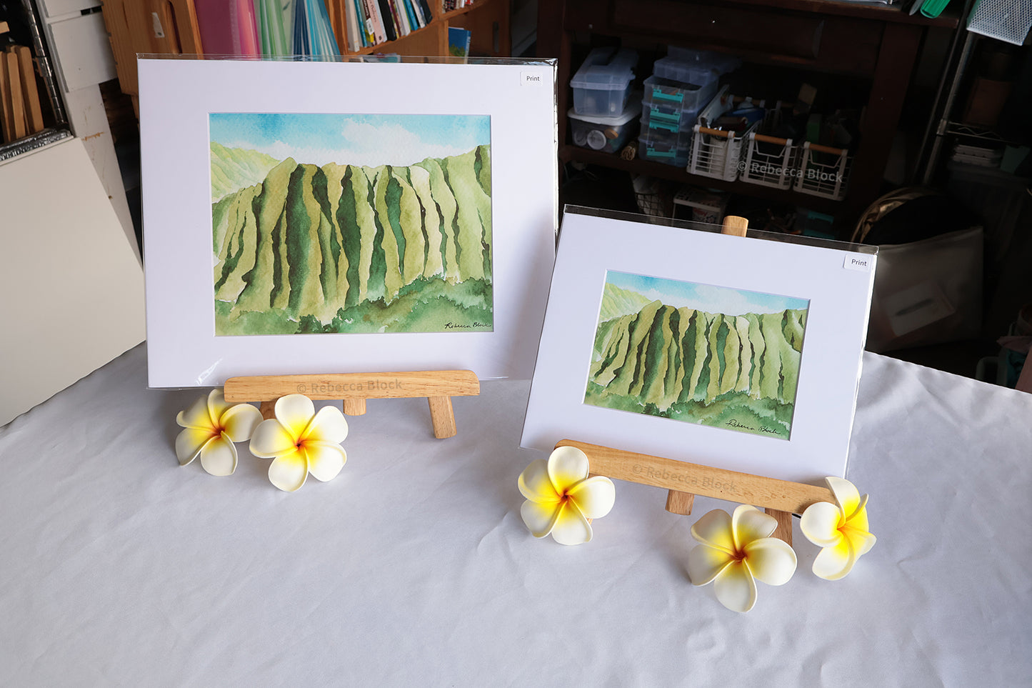 Two matted landscape art prints depicting green cliffs or dunes under a blue sky, displayed on small wooden easels on a white tablecloth with plumeria flowers in front, in an art studio setting.
