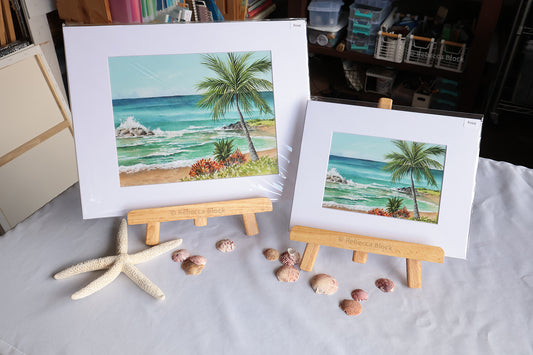 Two matted beach art prints displayed on small wooden easels on a white tablecloth, with a starfish and seashells arranged in front. The larger print reads 'Beach Breeze' and features turquoise ocean waves, a palm tree, sandy shore, and rocks.
