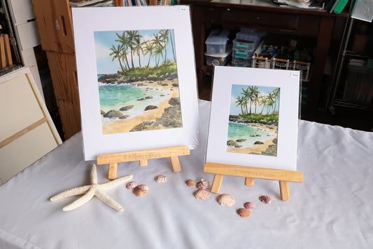 Two matted tropical beach paintings on wooden easels, displayed on a white cloth with seashells and a starfish in a home studio setting.