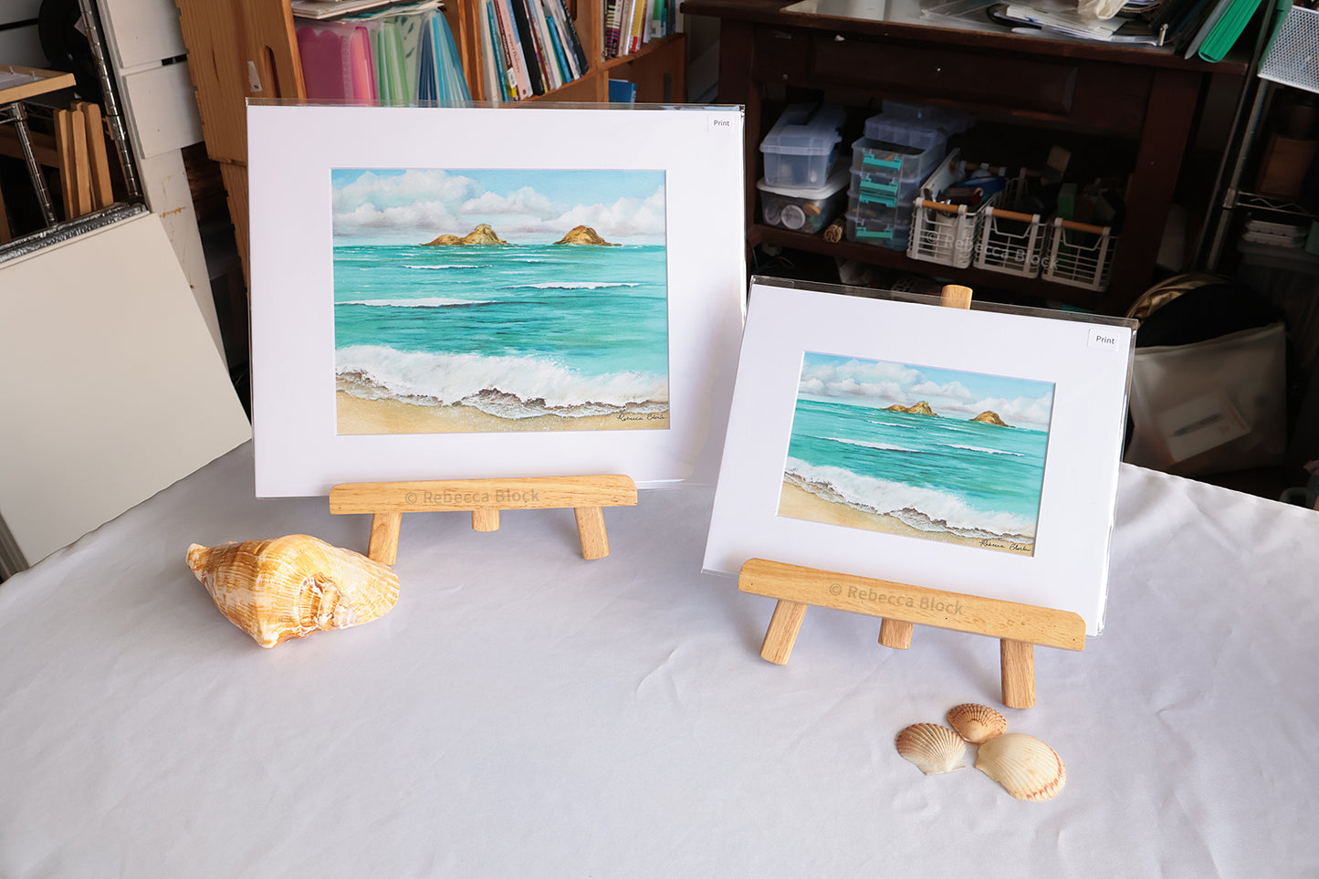 Two matted square paintings of a beach and sea scene with turquoise water, white waves, sandy shore, and small green islands, displayed on wooden easels on a white tablecloth. A large seashell sits beside the taller painting, and two smaller seashells are placed in front of the shorter painting.