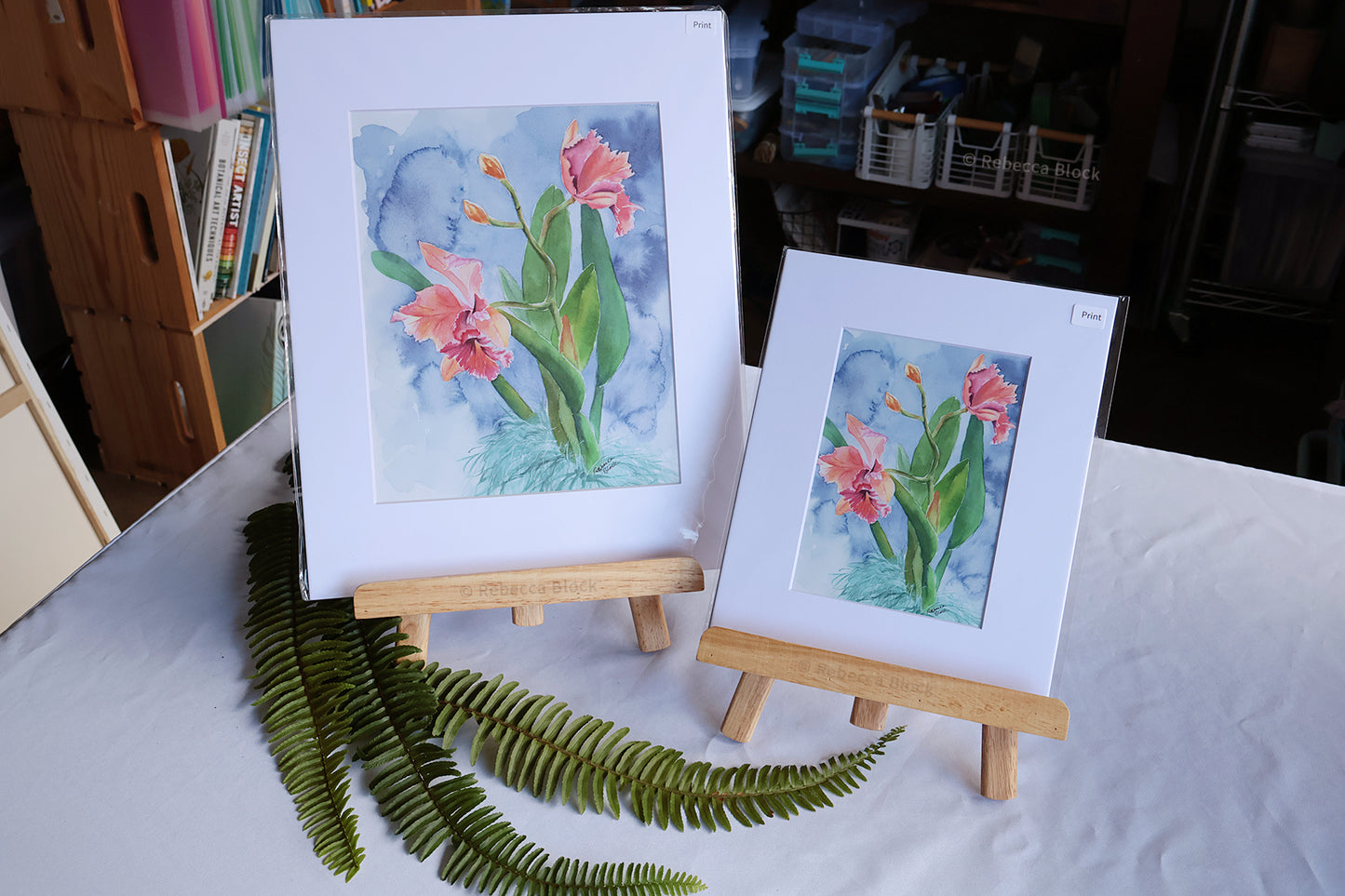 Two matted watercolor floral art prints on small wooden easels, displayed on a white tablecloth with green fern leaves in the foreground. The larger print stands upright and the smaller print leans against it.
