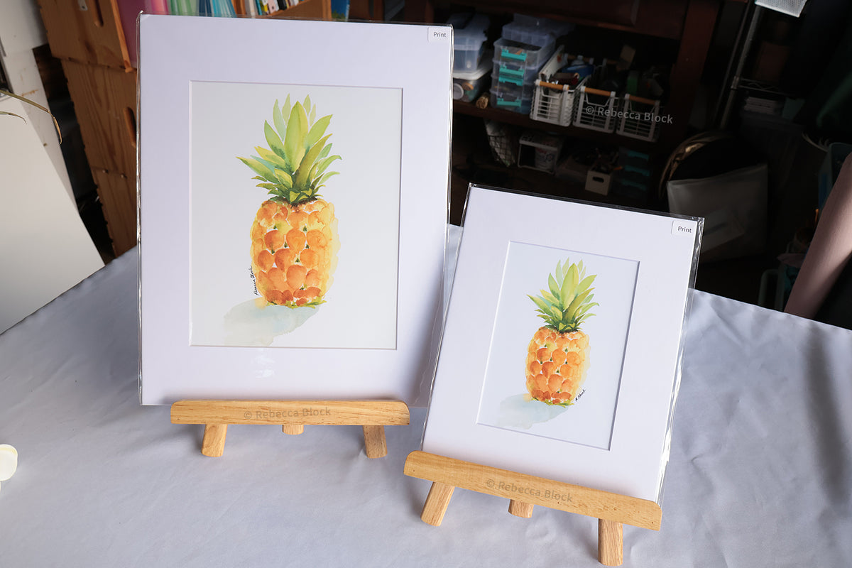Two matted watercolor pineapple art prints in different sizes, displayed on small wooden easels on a white tablecloth in a room setting.