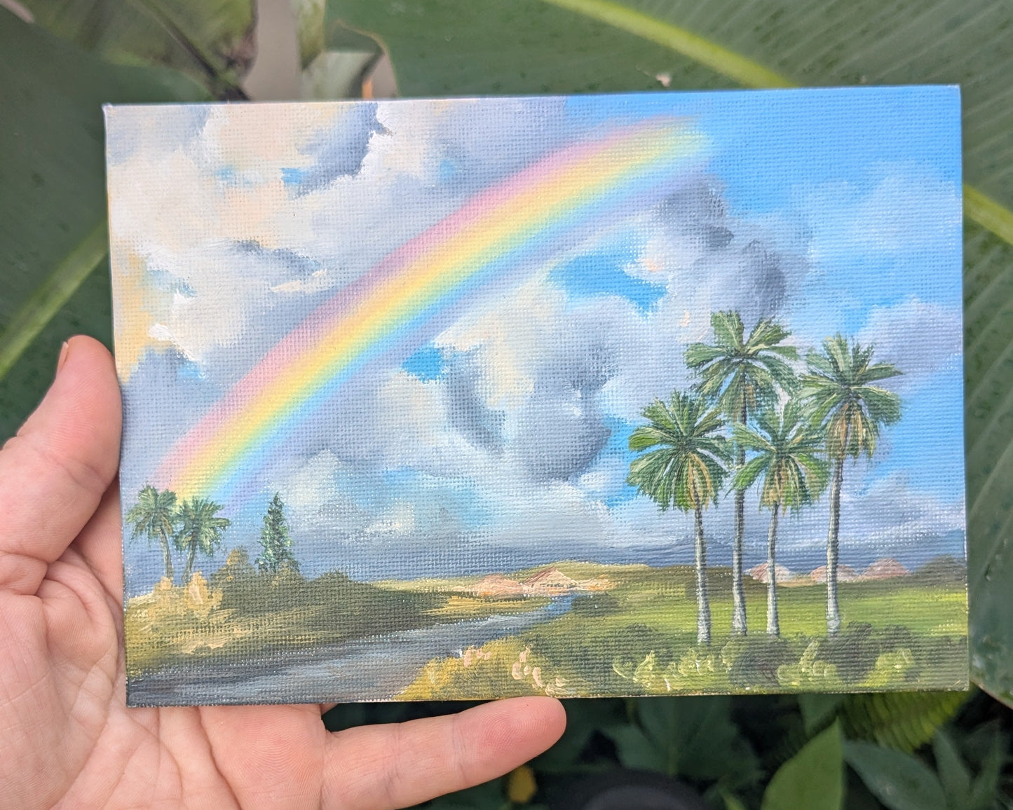 Hand holding a small painting of a rainbow and palm trees