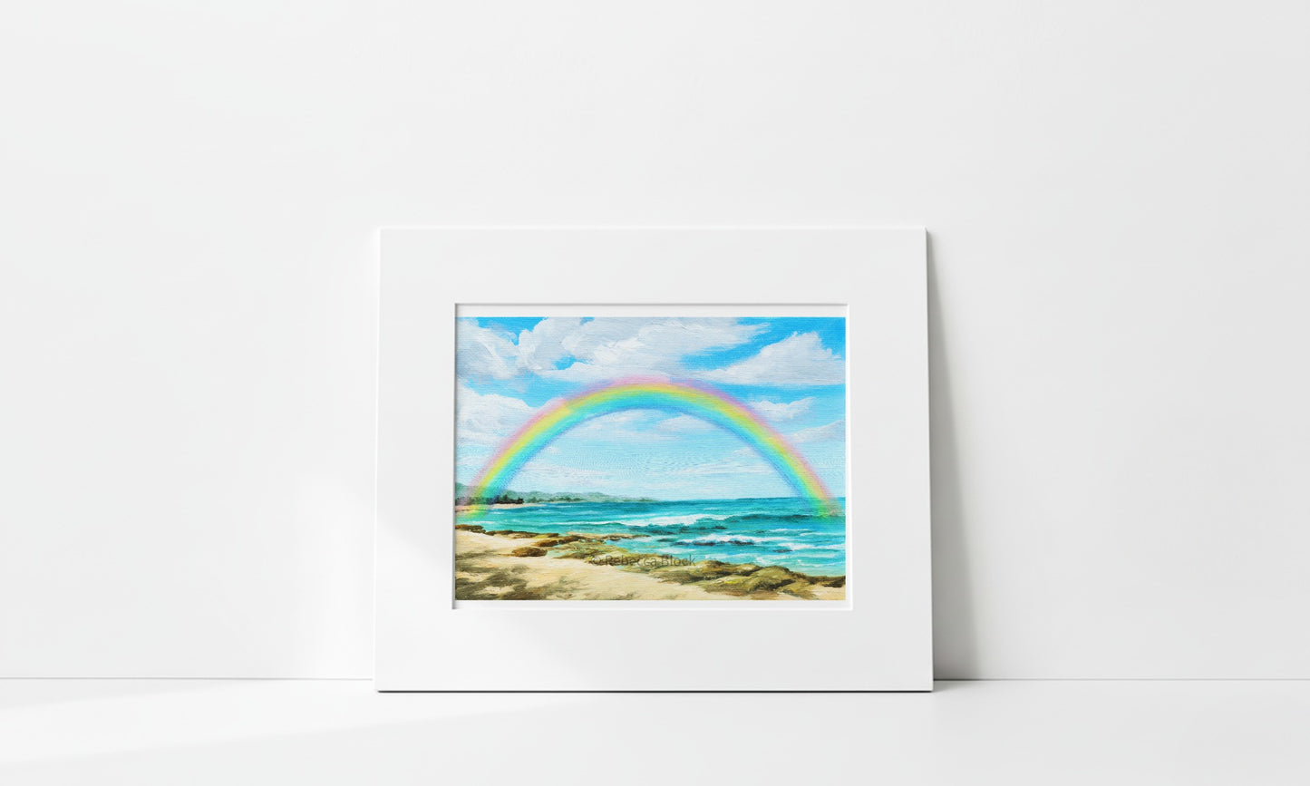 A matted art print depicting a vibrant rainbow arching over a sandy beach and turquoise ocean under a blue sky, displayed against a clean white wall.