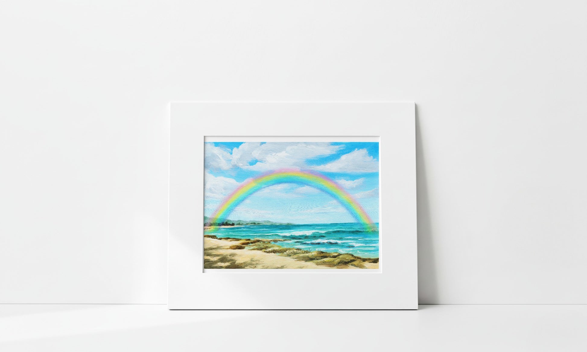A matted art print depicting a vibrant rainbow arching over a sandy beach and turquoise ocean under a blue sky, displayed against a clean white wall.