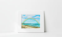 A matted art print depicting a vibrant rainbow arching over a sandy beach and turquoise ocean under a blue sky, displayed against a clean white wall.