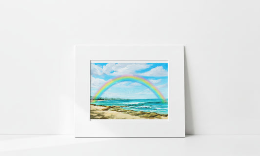 A matted art print depicting a vibrant rainbow arching over a sandy beach and turquoise ocean under a blue sky, displayed against a clean white wall.