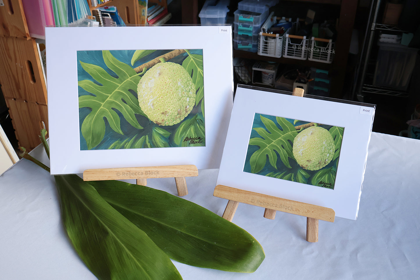Two matted botanical art prints depicting a green fruit among monstera leaves, displayed on small wooden easels on a white tablecloth with a large green leaf in the foreground.