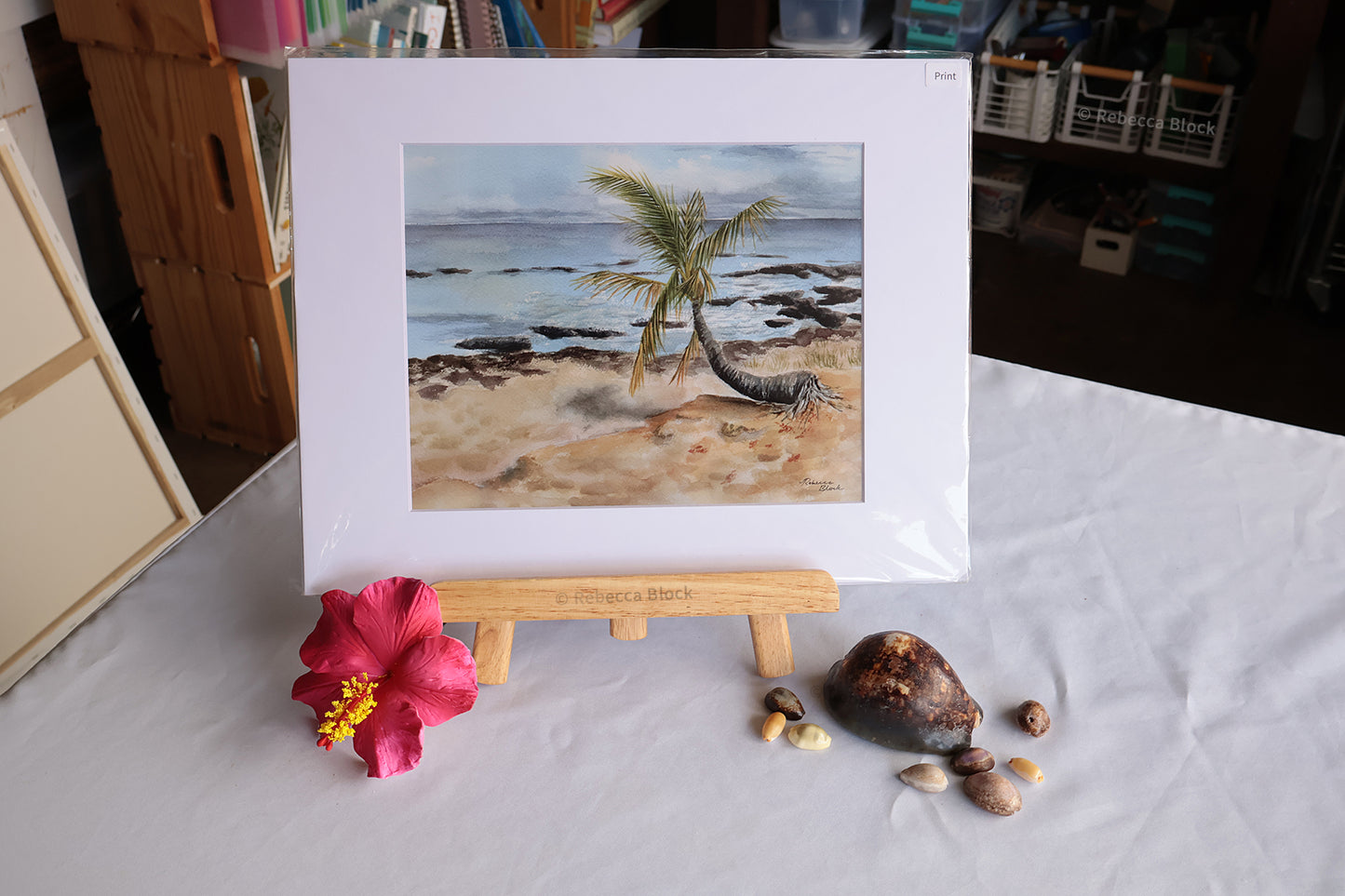 A matted beach painting displayed on a small wooden easel, with a pink hibiscus flower and seashells arranged on the table beside it. The room background is somewhat cluttered with shelves and household items.
