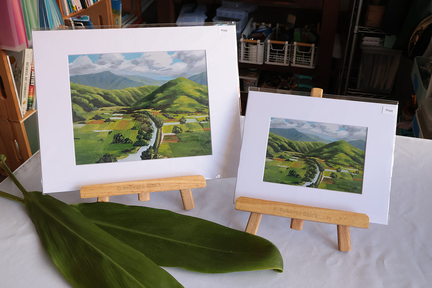 Two Hanalei, Hawaii landscape art prints displayed on wooden easels, featuring green rolling hills and a winding river under a cloudy sky, with a large green ti leaf in the foreground on a white tablecloth.