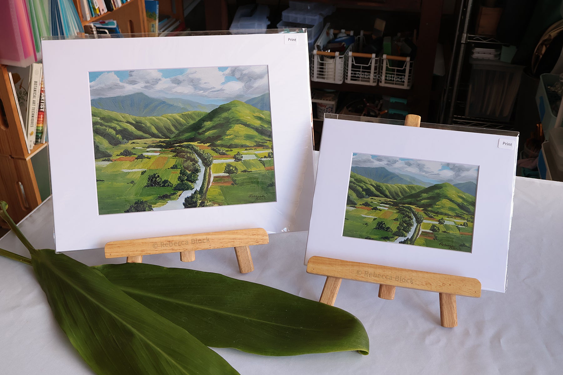 Two Hanalei, Hawaii landscape art prints displayed on wooden easels, featuring green rolling hills and a winding river under a cloudy sky, with a large green ti leaf in the foreground on a white tablecloth.