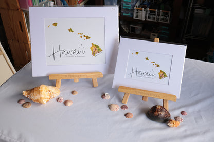Two framed art prints featuring a map of Hawaii with the word 'Hawaii' in script, displayed on small wooden easels on a white tablecloth with seashells around them.