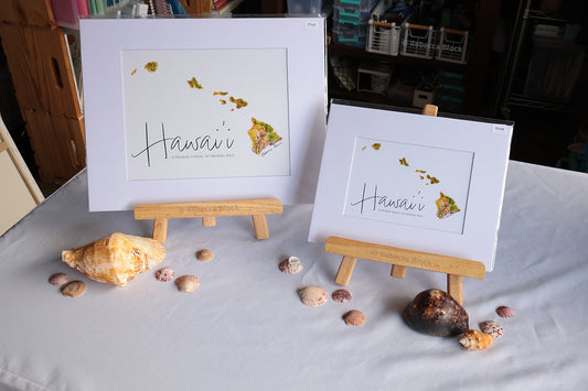 Two framed art prints featuring a map of Hawaii with the word 'Hawaii' in script, displayed on small wooden easels on a white tablecloth with seashells around them.