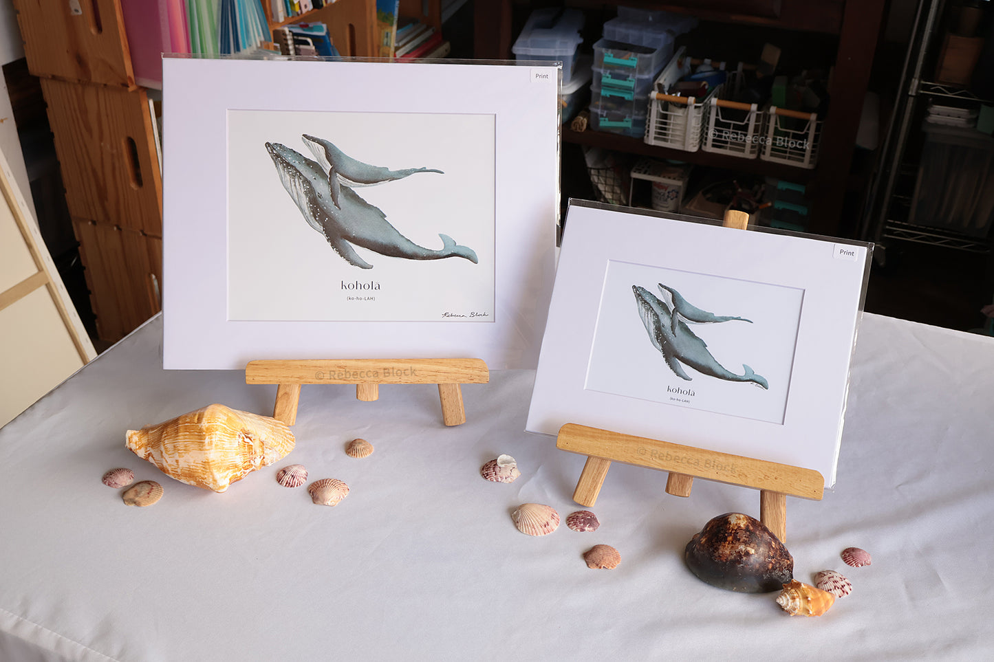 Two whale art prints displayed on small wooden easels on a white tablecloth, surrounded by seashells. The background shows a shelf with books and storage items.