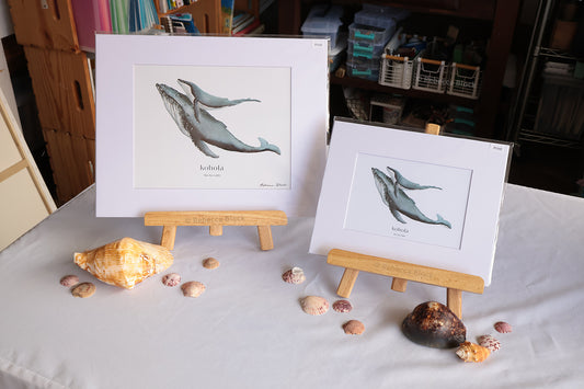 Two whale art prints displayed on small wooden easels on a white tablecloth, surrounded by seashells. The background shows a shelf with books and storage items.