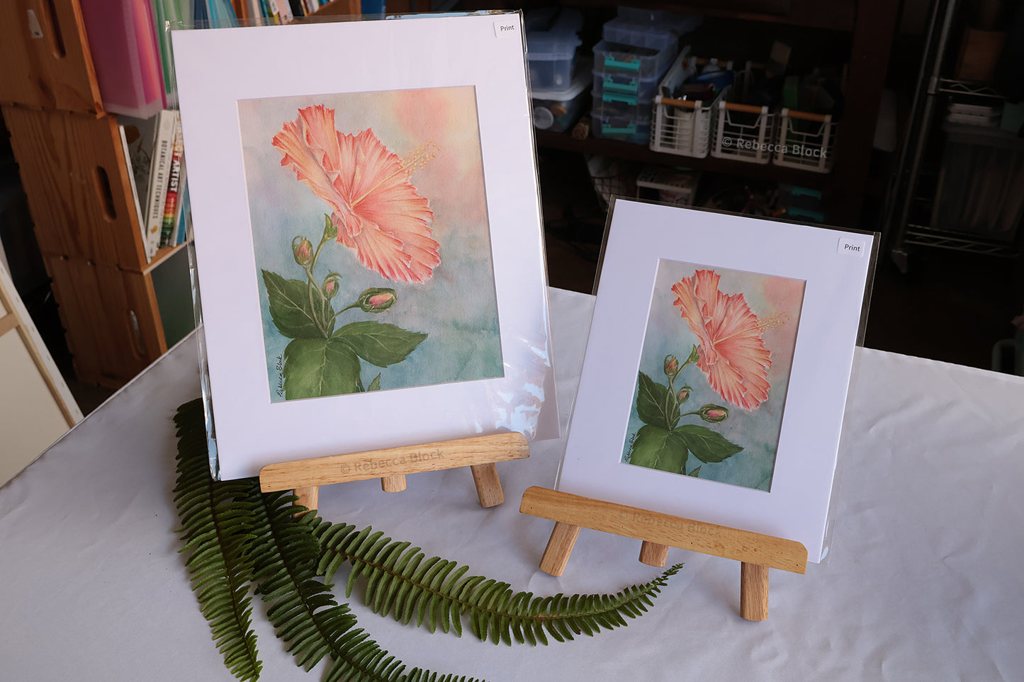 Two matted floral art prints displayed on small wooden easels on a white tablecloth-covered surface, with green fern leaves in the foreground and a real indoor room visible in the background.