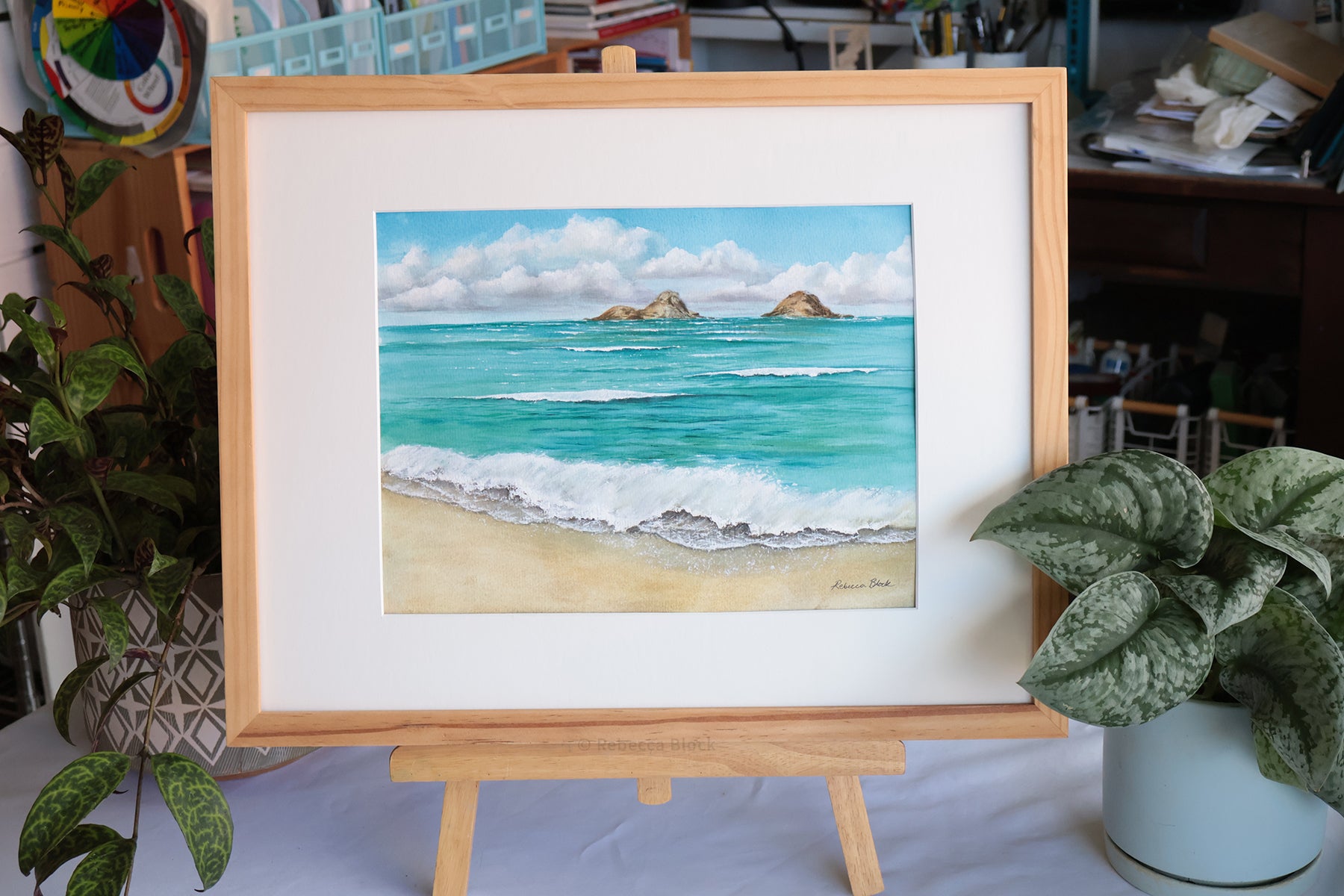A framed watercolor painting of a beach and ocean scene displayed on a wooden easel. The artwork features turquoise water, white waves, sandy shore, and distant rocky islands under a blue sky with clouds.