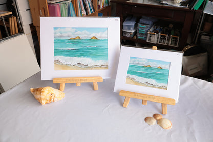 Two matted square paintings of a beach and sea scene with turquoise water, white waves, sandy shore, and small green islands, displayed on wooden easels on a white tablecloth. A large seashell sits beside the taller painting, and two smaller seashells are placed in front of the shorter painting.
