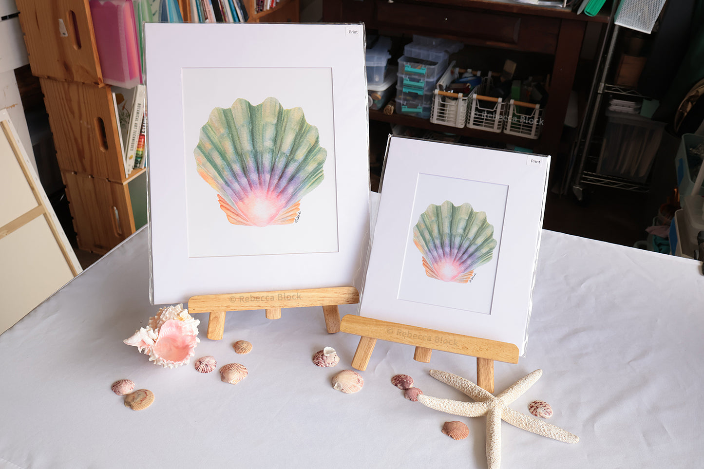 Two matted seashell art prints displayed on small wooden easels on a white tablecloth, with seashells and a starfish arranged around them in a room setting.