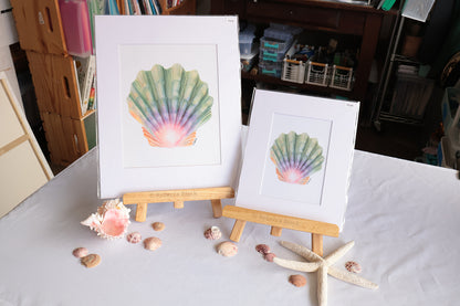 Two matted seashell art prints displayed on small wooden easels on a white tablecloth, with seashells and a starfish arranged around them in a room setting.