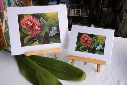 Two framed floral art prints displayed on small wooden easels on a white tablecloth, with a large green leaf in the foreground. The artwork depicts bright red flowers with green leaves and small blue blossoms.