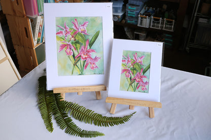 Two matted art prints featuring pink orchids on wooden easels, displayed on a white tablecloth with green fern leaves in the foreground. The background shows a workshop or studio setting.