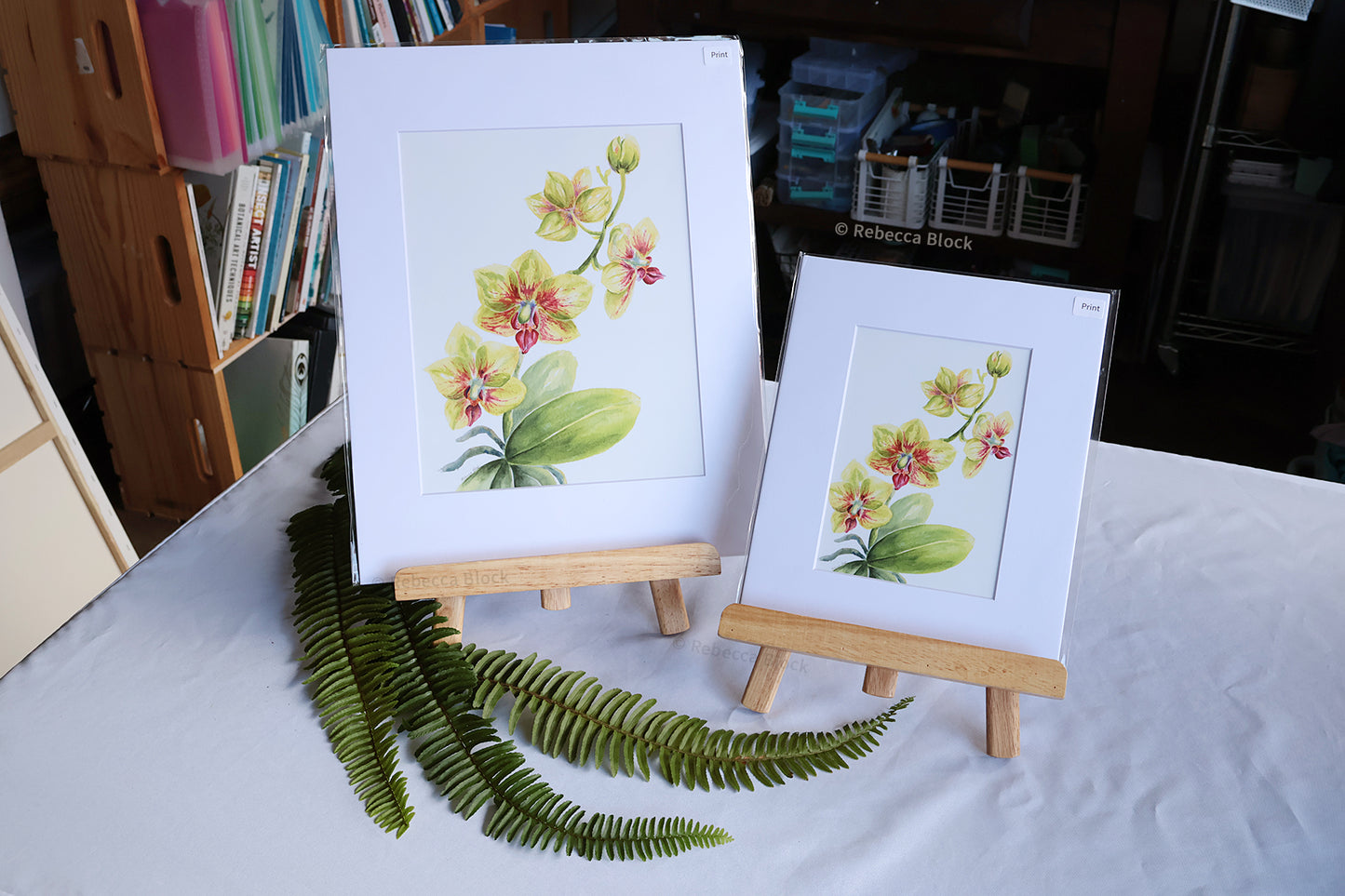 Two matted floral art prints with white mats are displayed on small wooden easels on a white tablecloth. The larger print shows yellow and pink orchids with green leaves, and the smaller print shows a similar floral design.