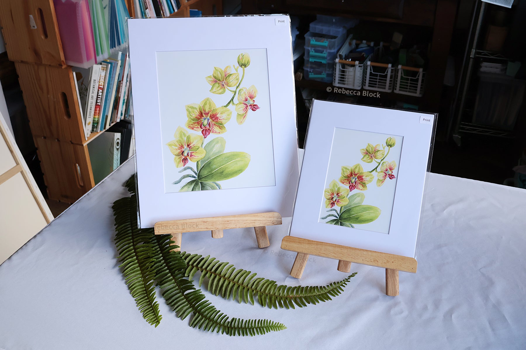 Two matted floral art prints with white mats are displayed on small wooden easels on a white tablecloth. The larger print shows yellow and pink orchids with green leaves, and the smaller print shows a similar floral design.