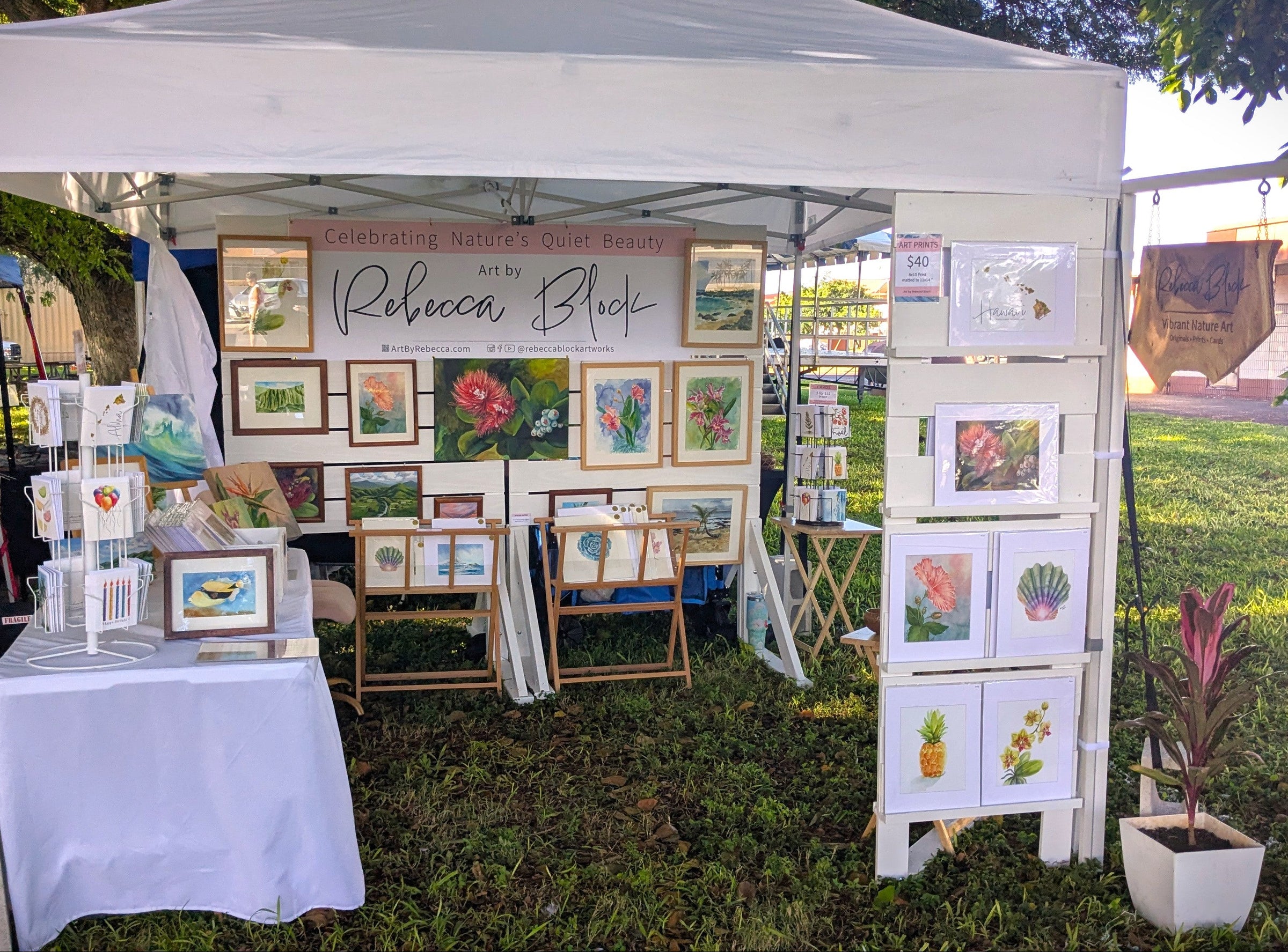 Artist's booth with nature, tropical and Hawai'i themed artwork displayed outdoors on a grassy area