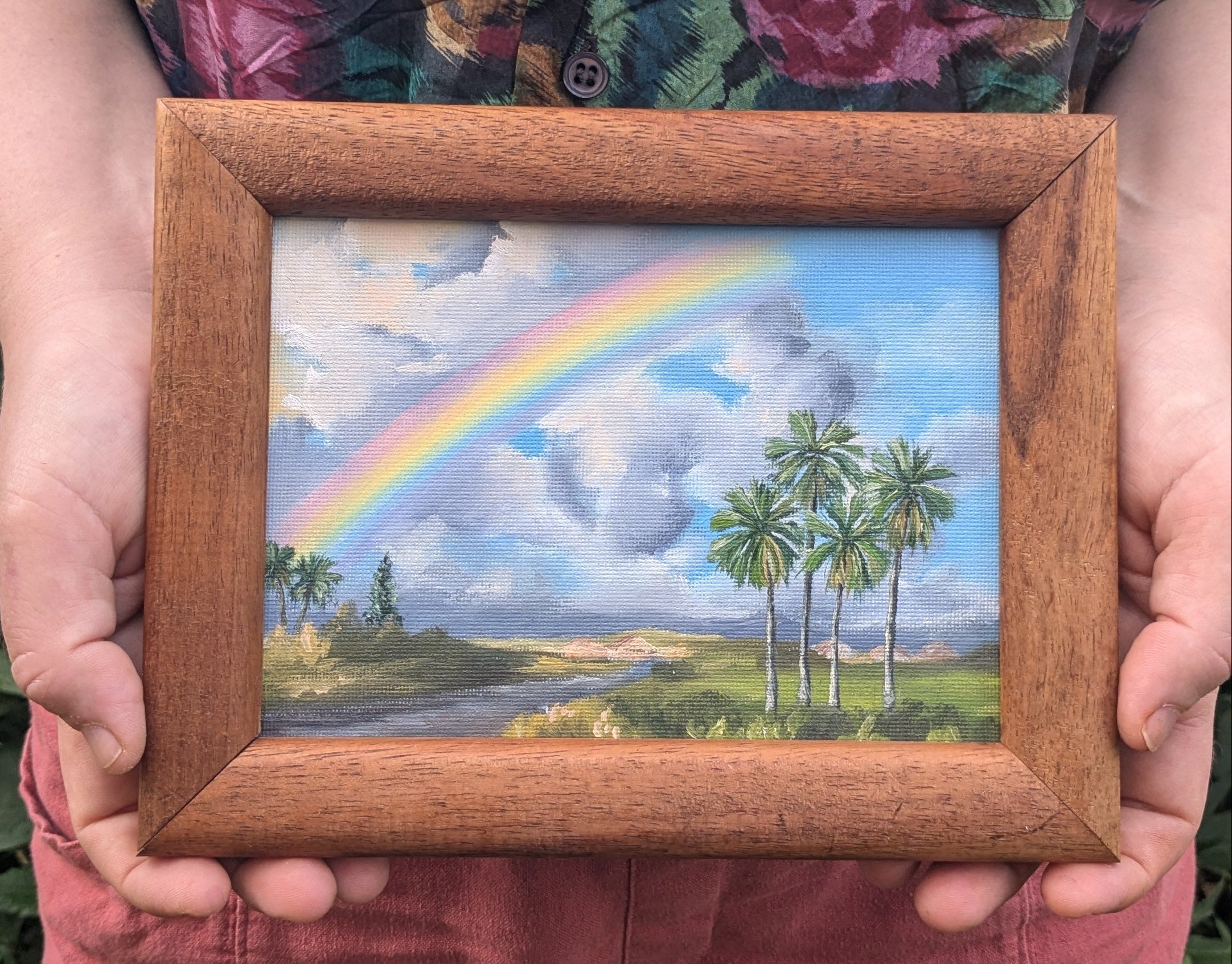 Person holding a framed painting of a rainbow and palm trees